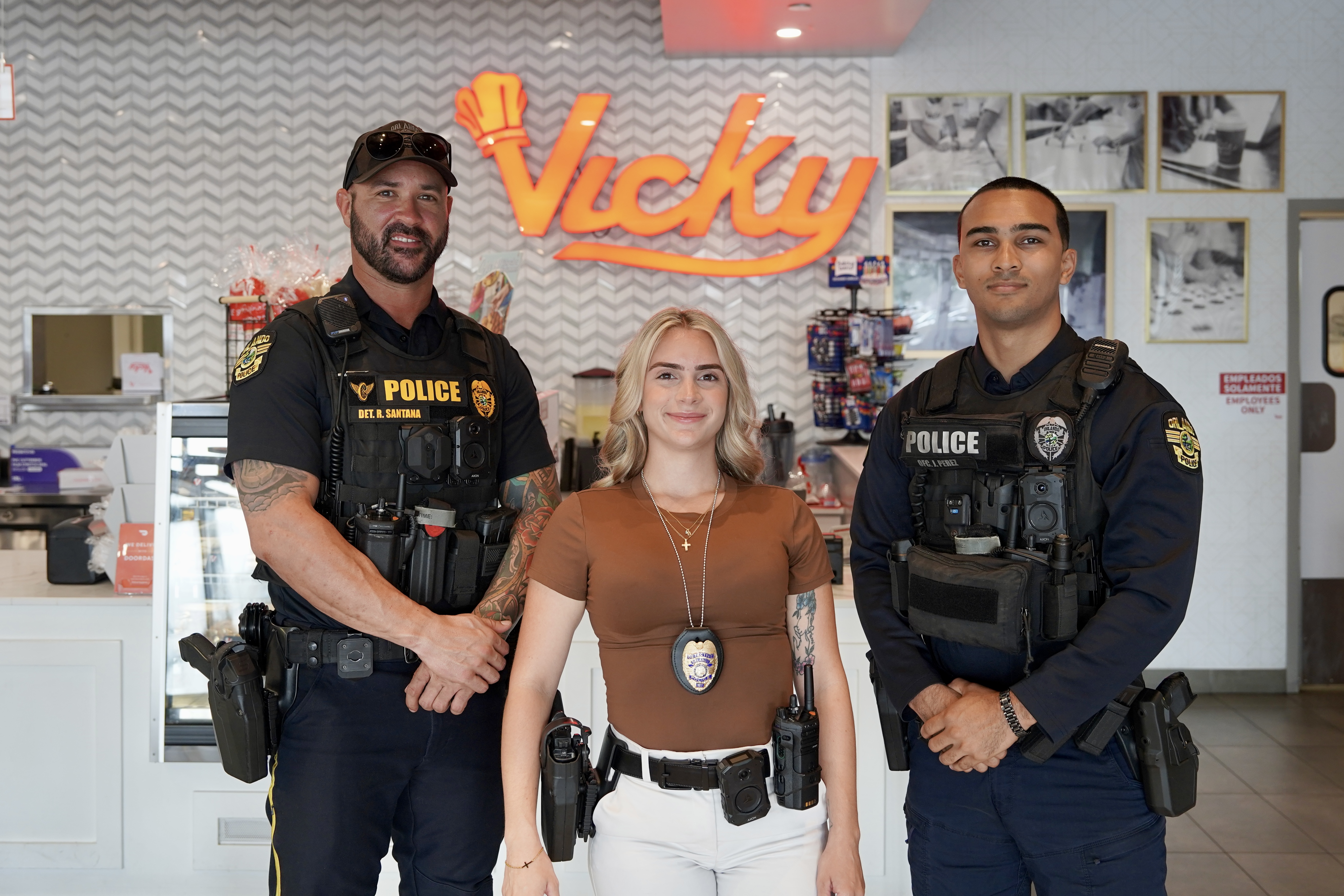 Three OPD police officers stand inside Vicky’s Bakery with a “Vicky” sign behind them. Two wear dark police vests, and the officer in the center wears a badge and utility belt. The trio stands in front of a counter and display shelves, posing for a photo.
