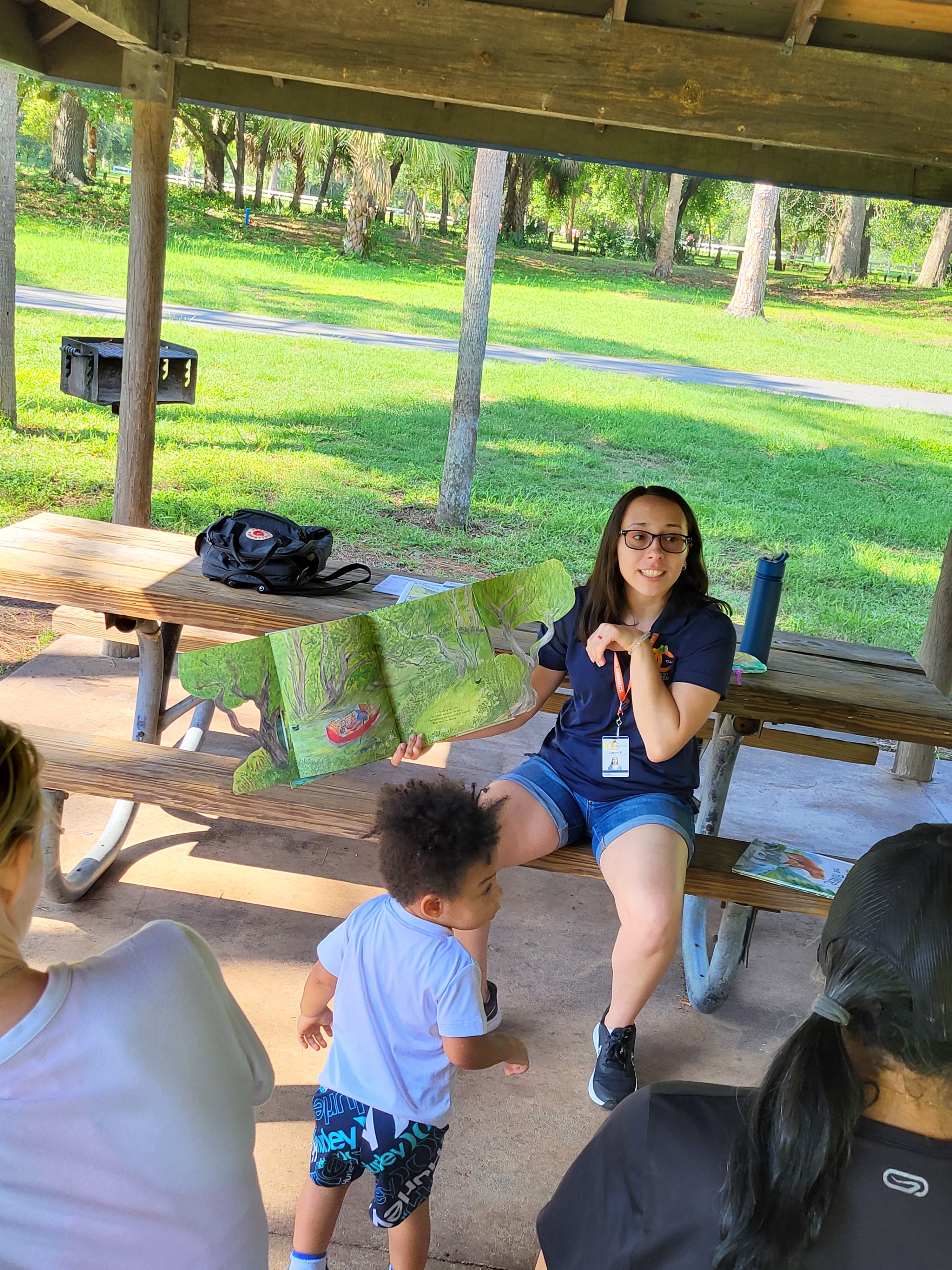 Woman sitting on a picnic table in a park pavilion reading a book to children