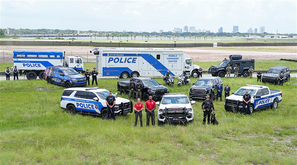 A group of Orlando Police Department personnel stand in a grassy field with a variety of police vehicles, including SUVs, trucks, motorcycles, an armored vehicle, and a large mobile command center. The city skyline is visible in the background.